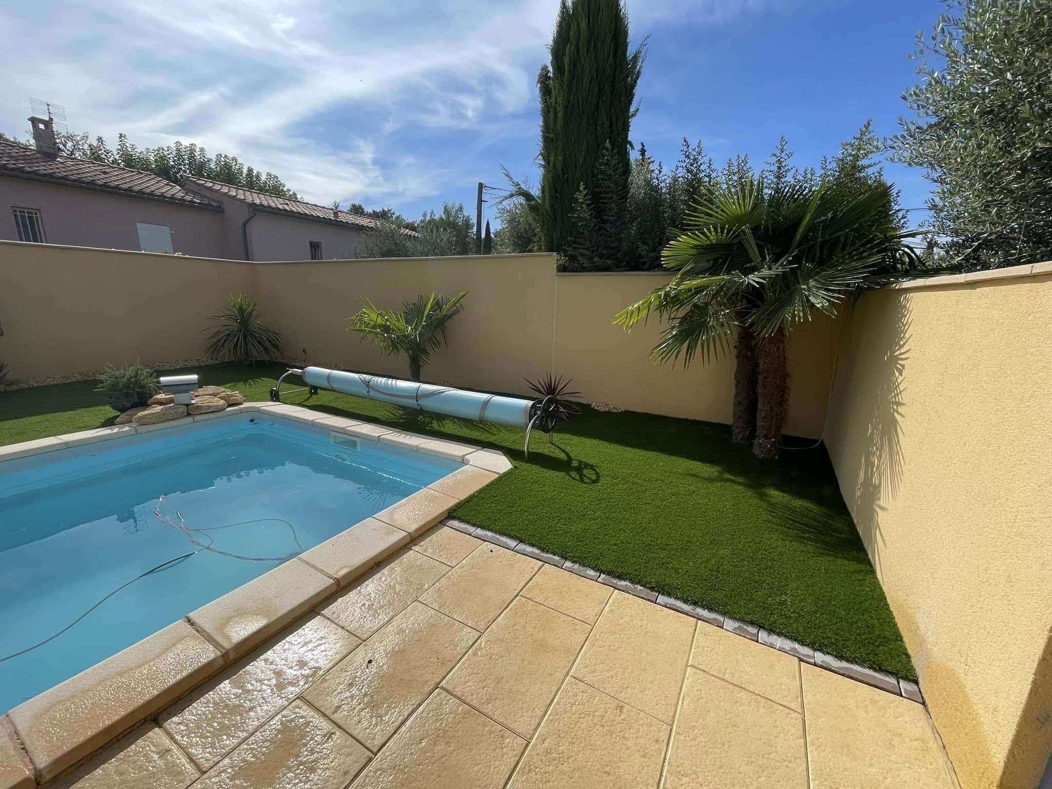 Aménagement piscine avec gazon synthétique et terrasse Piscine moderne avec gazon synthétique, palmiers et dallage beige dans un jardin ensoleillé clos de murs.