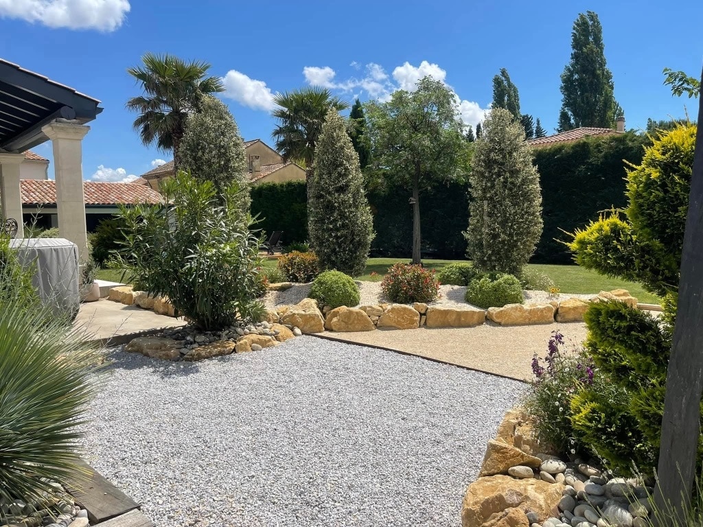 Jardin paysager avec gravier blanc, rochers décoratifs, palmiers et arbustes sous un ciel bleu ensoleillé.